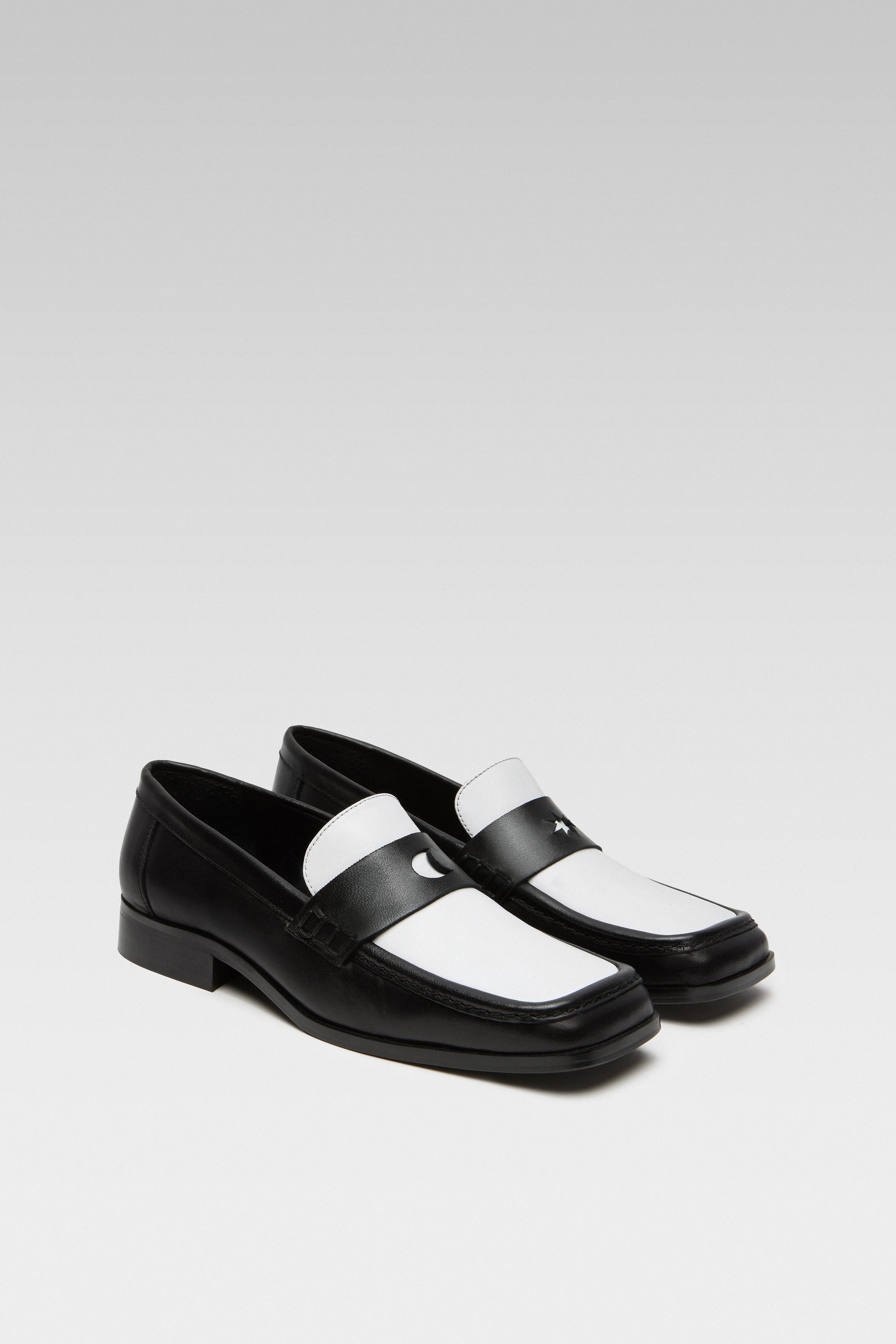 Three-quarter view of EDLA Earth black-and-white penny loafers for women crafted from leather with a square toe and star and moon inserts at the front.