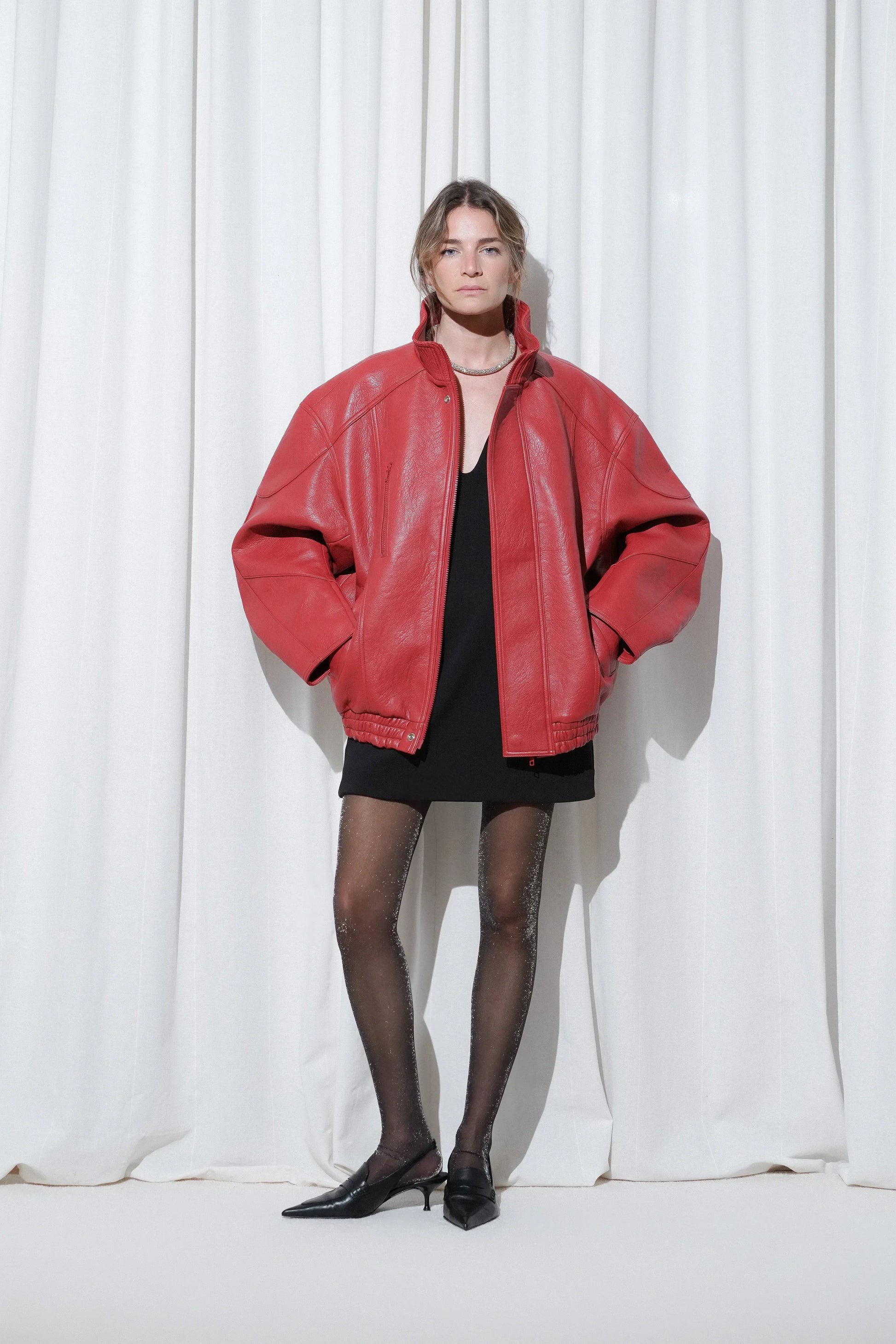 Model styled in a bold red oversized leather jacket layered over a black mini dress, paired with EDLA Cosmic Lurex Silver Shimmer Tights and EDLA Uranus Black Slingback Heels for a refined evening statement look.
