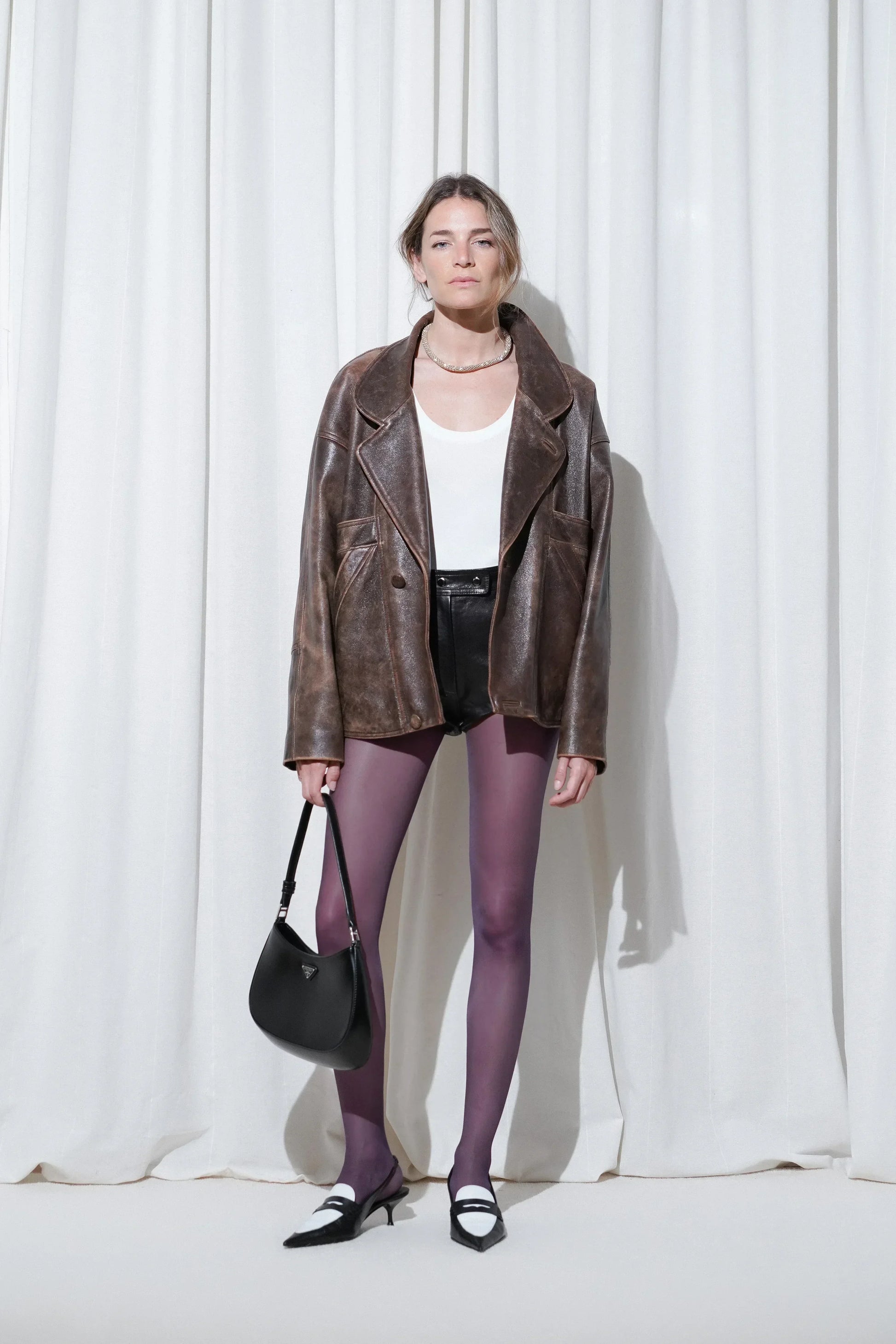 Model wearing a night-out look with an oversized brown leather jacket, white tank top, black mini shorts, EDLA Galaxy Purple Tights, and EDLA Uranus Black & White Slingbacks.