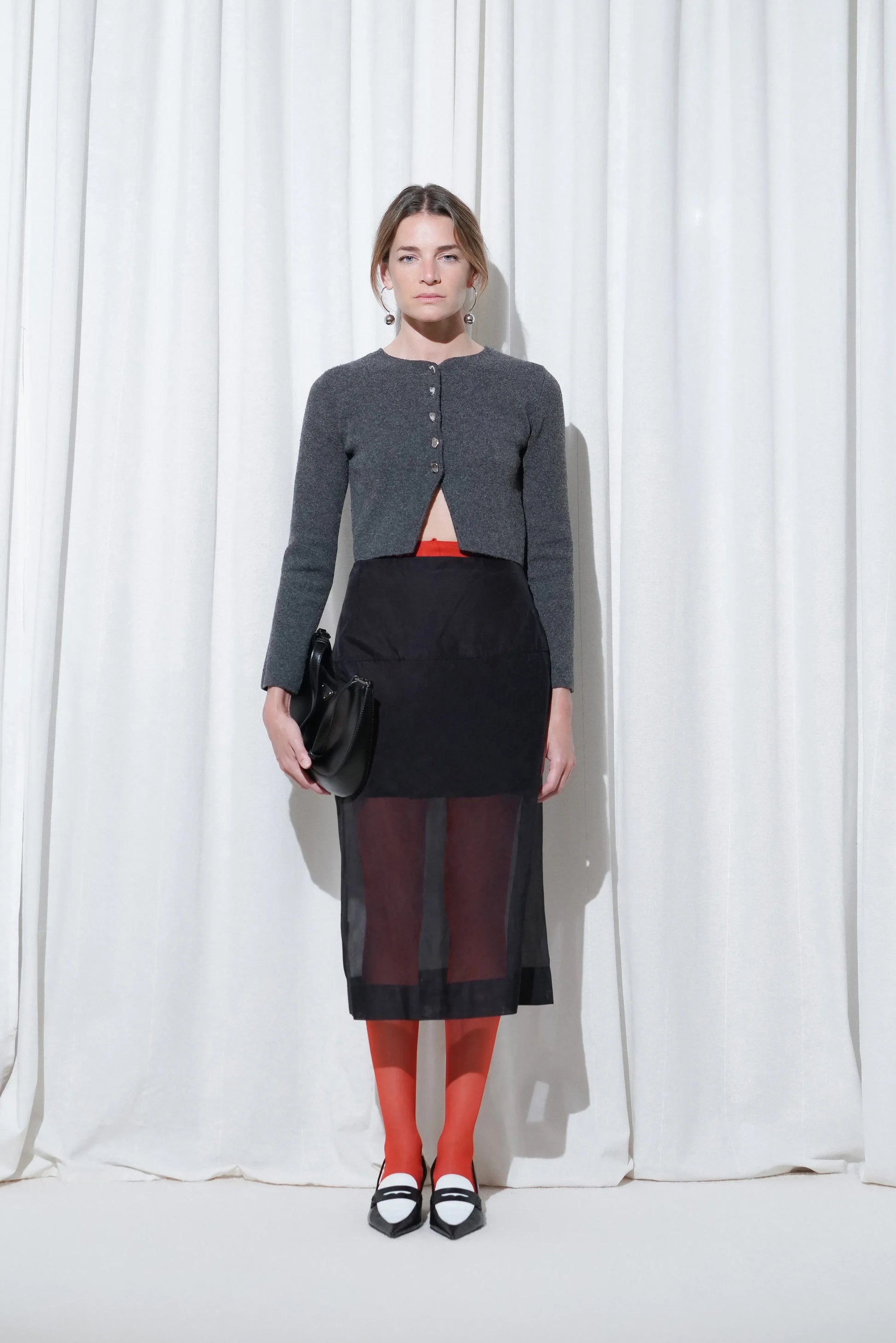Model wearing an evening outfit with a cropped grey cardigan, sheer black midi skirt, EDLA Galaxy Red Tights and EDLA Uranus Black and White Slingbacks.