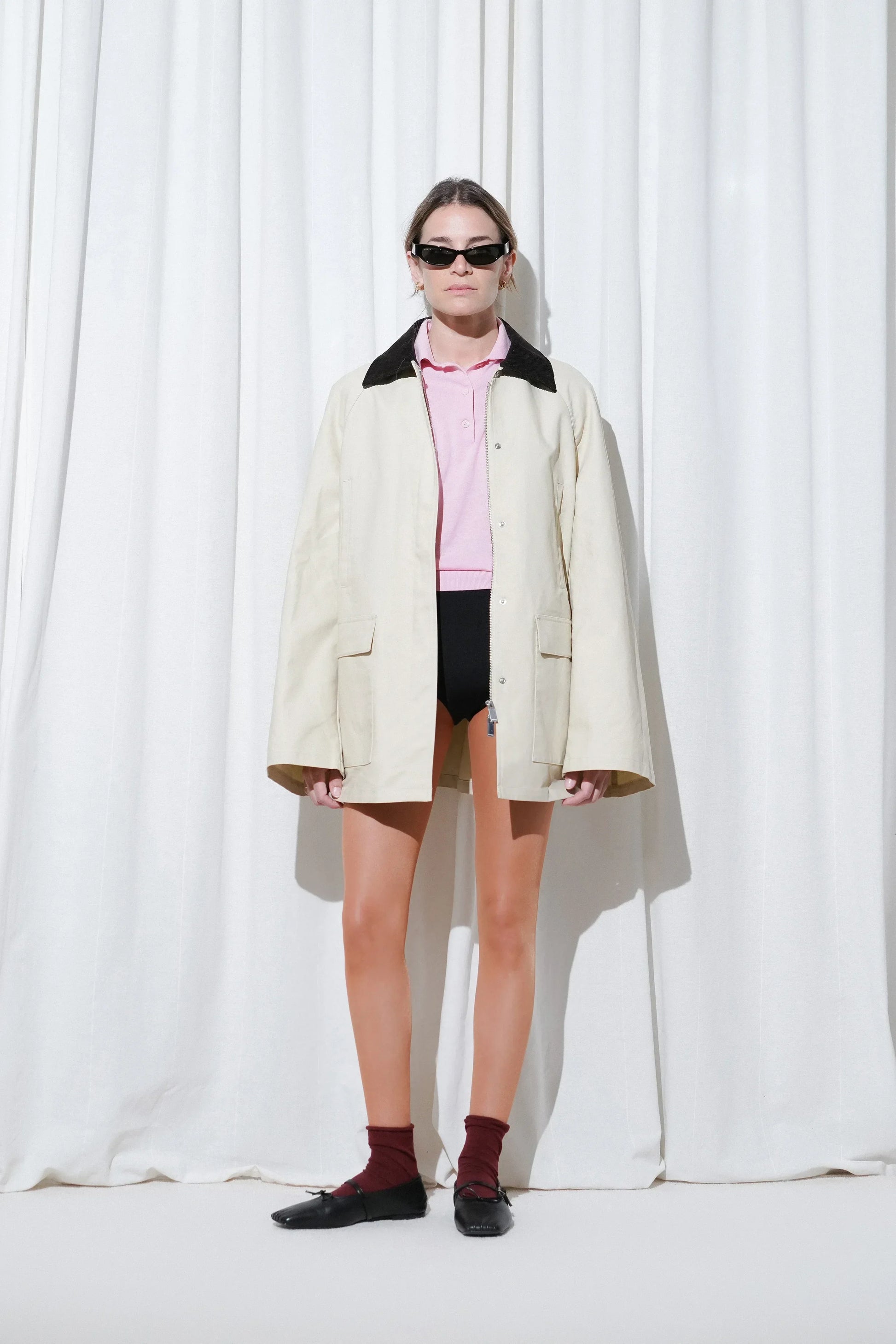 Model in a preppy casual look with a cream oversized jacket layered over a pink polo shirt and black shorts, styled with EDLA Burgundy Ursa Socks and EDLA Mars Black Leather Ballet Flats.
