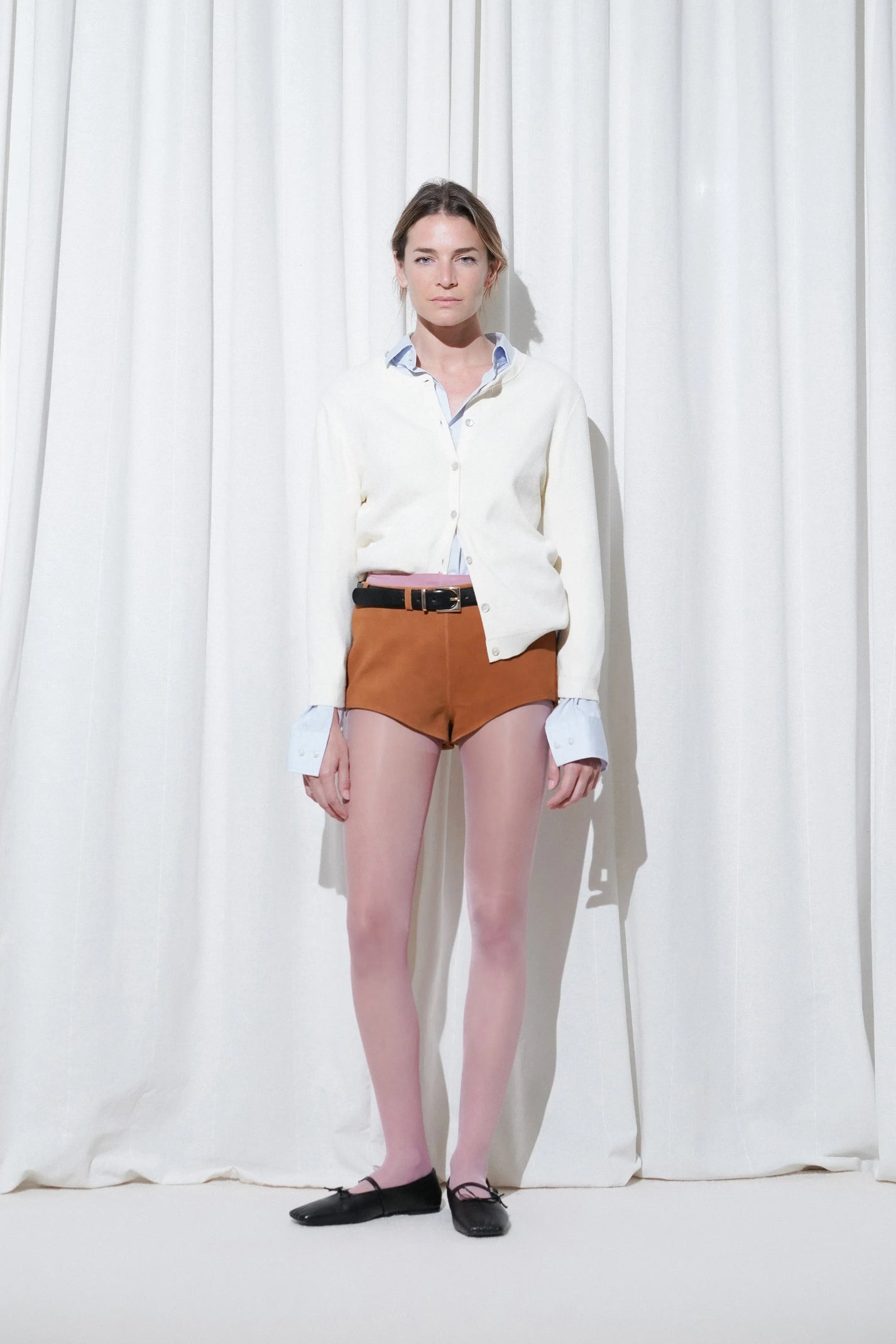 Model wearing a casual chic outfit with a light blue button-down shirt layered under a cream cardigan, brown suede shorts, EDLA Galaxy Pink Tights, and EDLA Mars Black Ballet Flats.