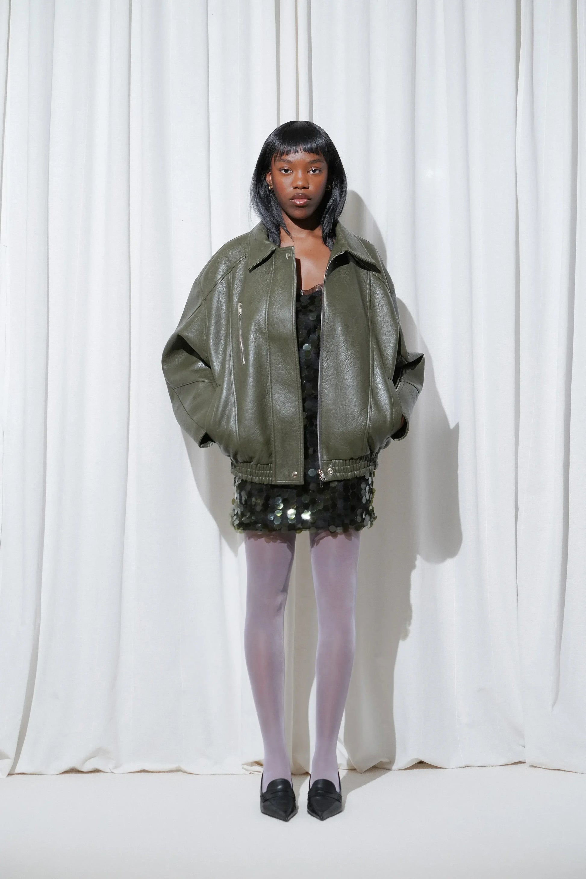 A model wearing a party look with khaki sequin mini dress layered with oversized leather jacket, EDLA Galaxy Lilac Tights, and EDLA Uranus Black Slingbacks.