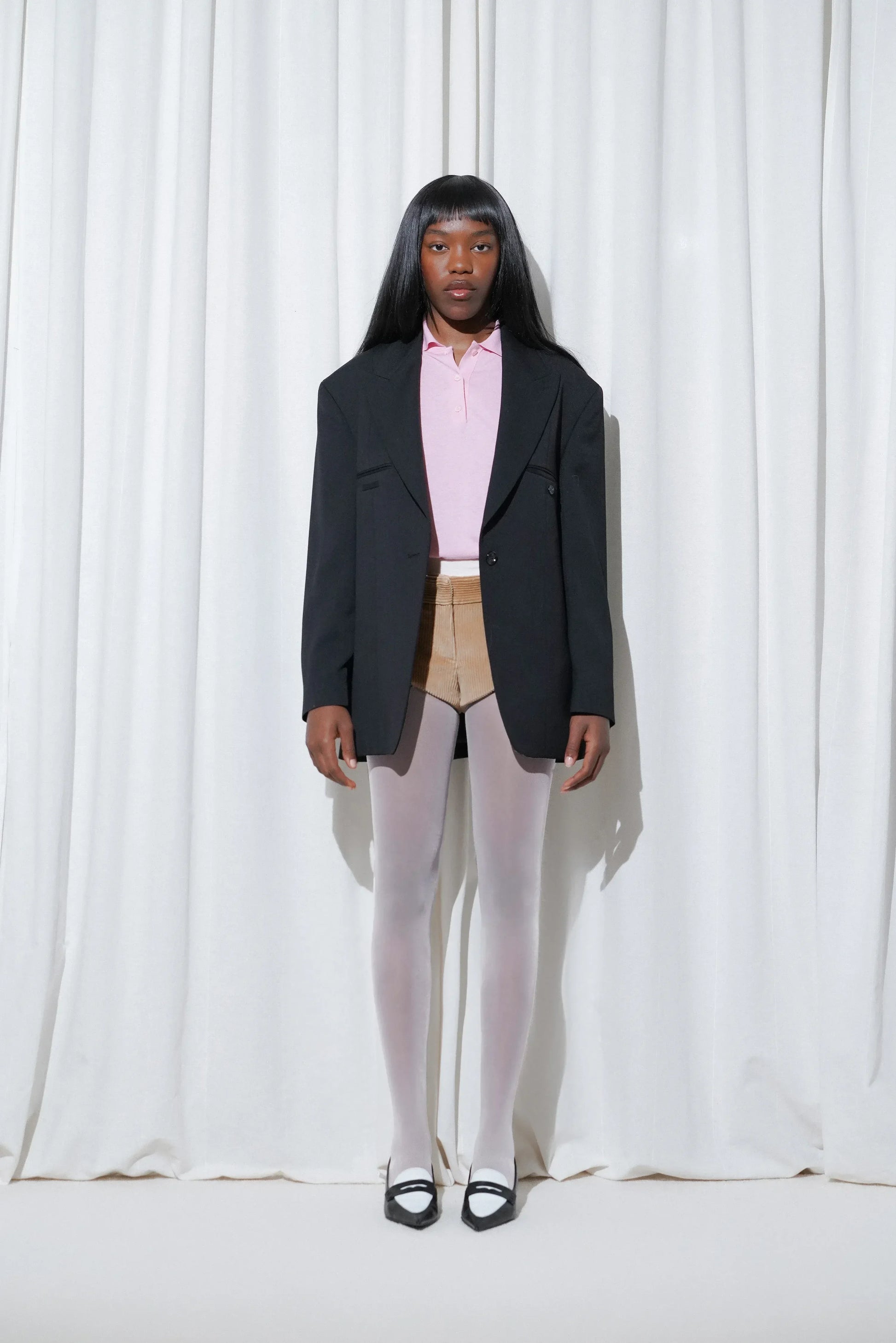 Model wearing a smart-casual business outfit with a pink polo sweater layered under an oversized black blazer, beige tailored shorts, EDLA Galaxy Light Grey Tights, and EDLA Uranus Black and White Slingbacks.