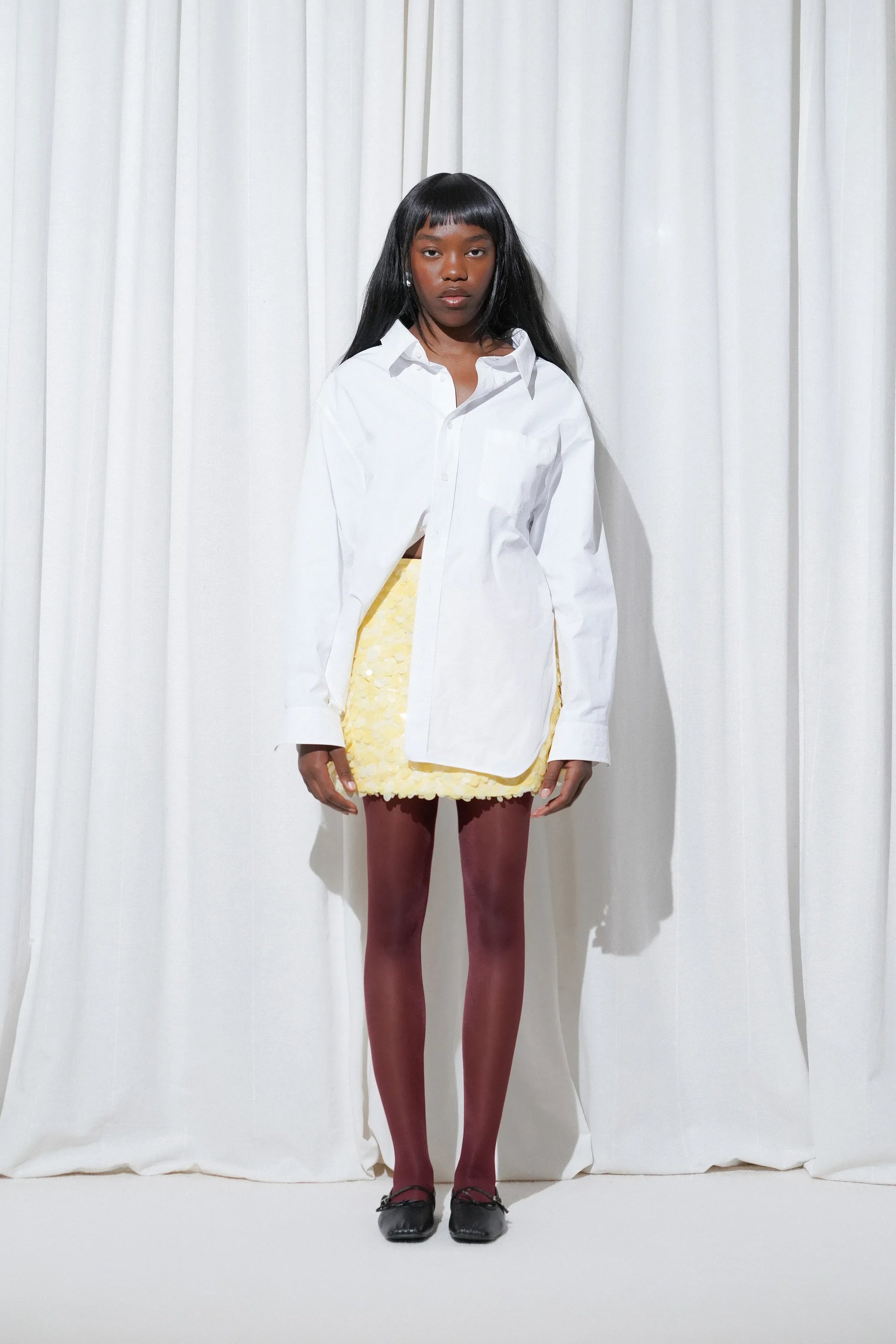 Model wearing a dinner outfit with an oversized crisp white shirt, a yellow sequins mini skirt, EDLA Galaxy Burgundy Tights, and EDLA Mars Black Ballet Flats.