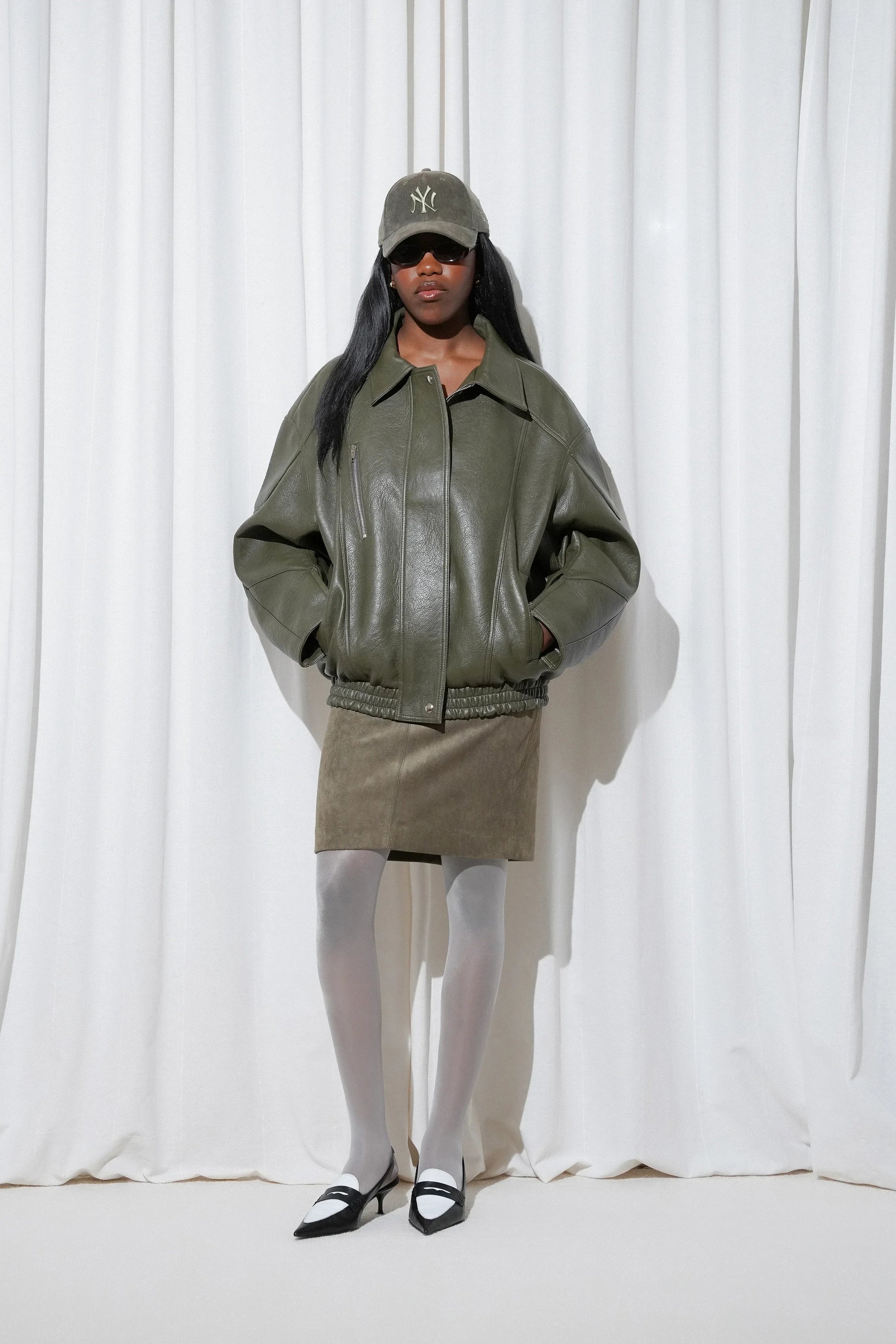 Model wearing a sporty chic outfit with a matching olive green cap, oversized leather jacket, and mini skirt, styled with EDLA Galaxy Light Grey Tights and EDLA Uranus Black and White Slingbacks.