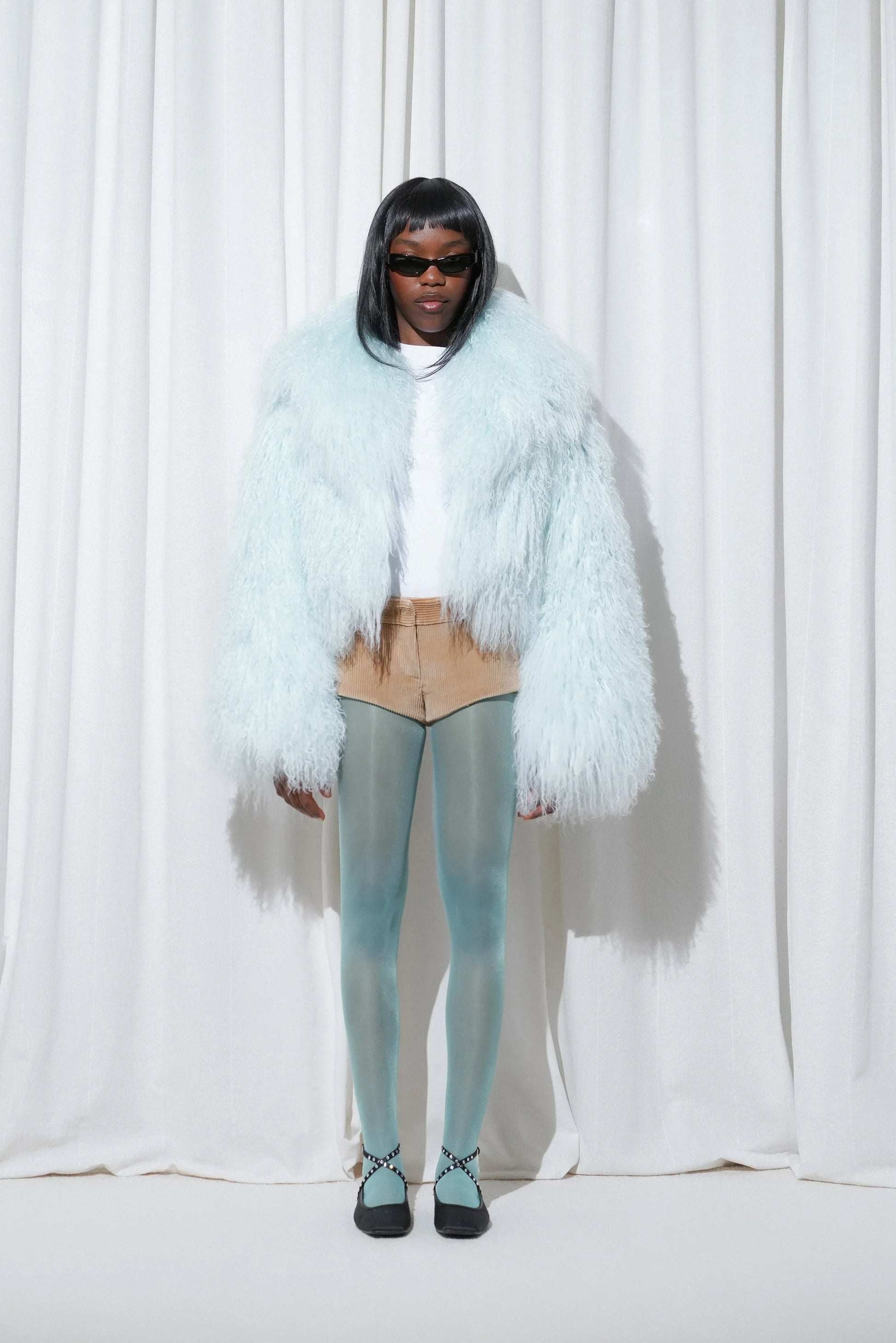 Model wearing a statement nightout look with a baby blue faux fur jacket, white top, beige shorts, EDLA Galaxy Aquamarine Tights, and EDLA Saturn Black Ballet Flats.