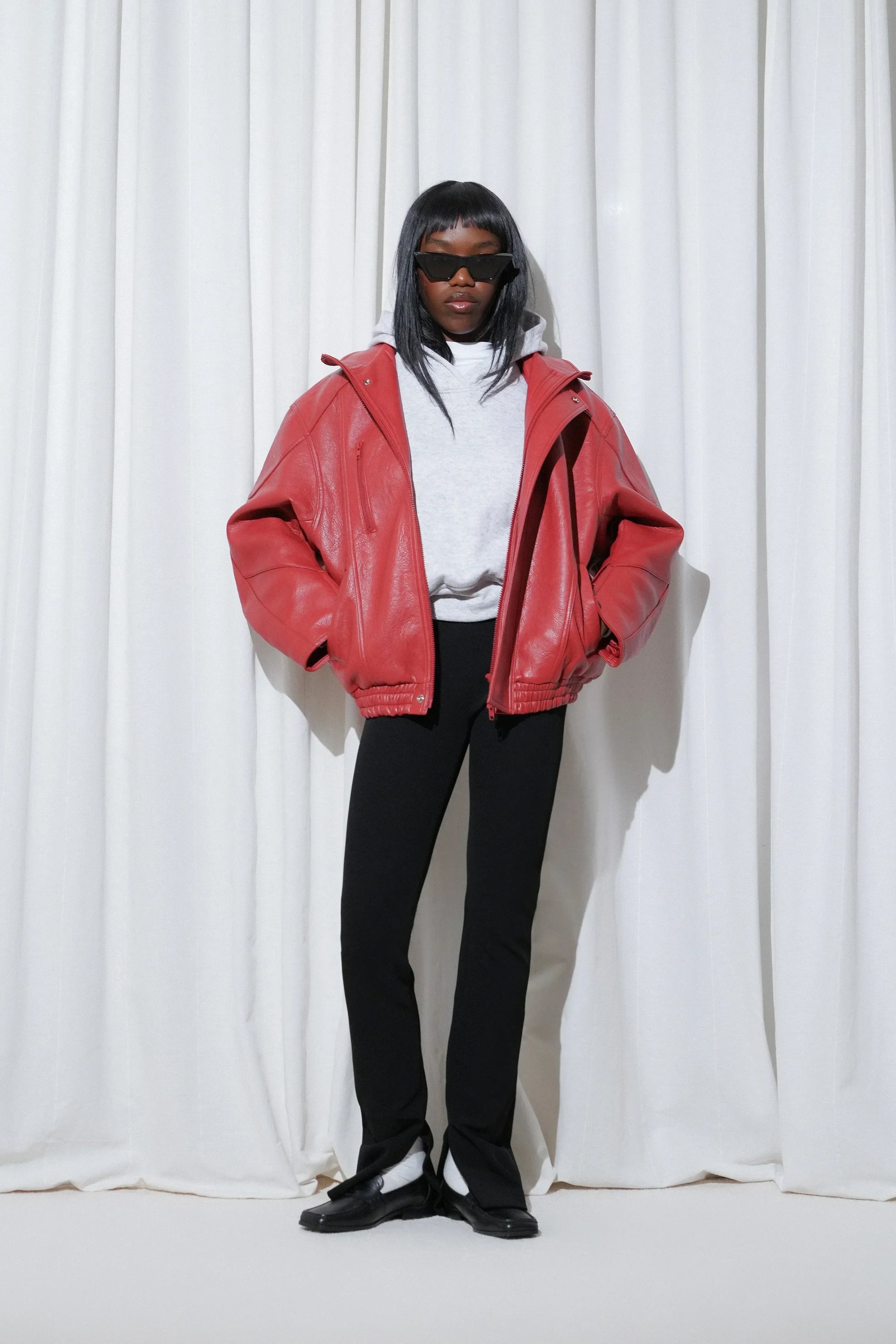 Model in airport outfit with oversized red leather jacket, grey sweatshirt, black slim trousers, EDLA Ursa White Socks and EDLA Earth Black Loafers.