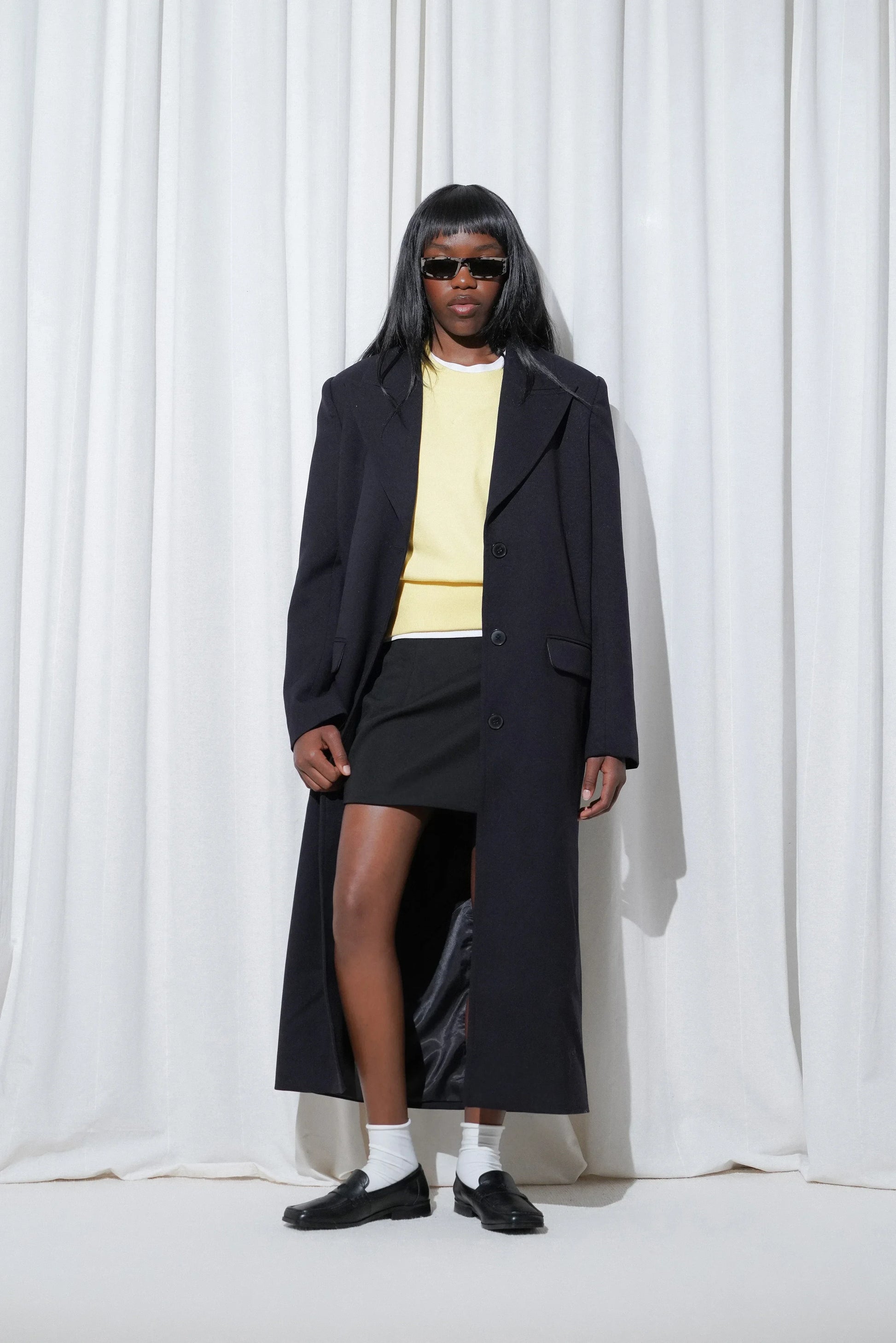 Model in a sleek urban outfit featuring a long navy coat, pastel yellow sweater layered over a white tee, black mini skirt, EDLA Ursa White Socks, and EDLA Black Earth Loafers, styled with narrow sunglasses.