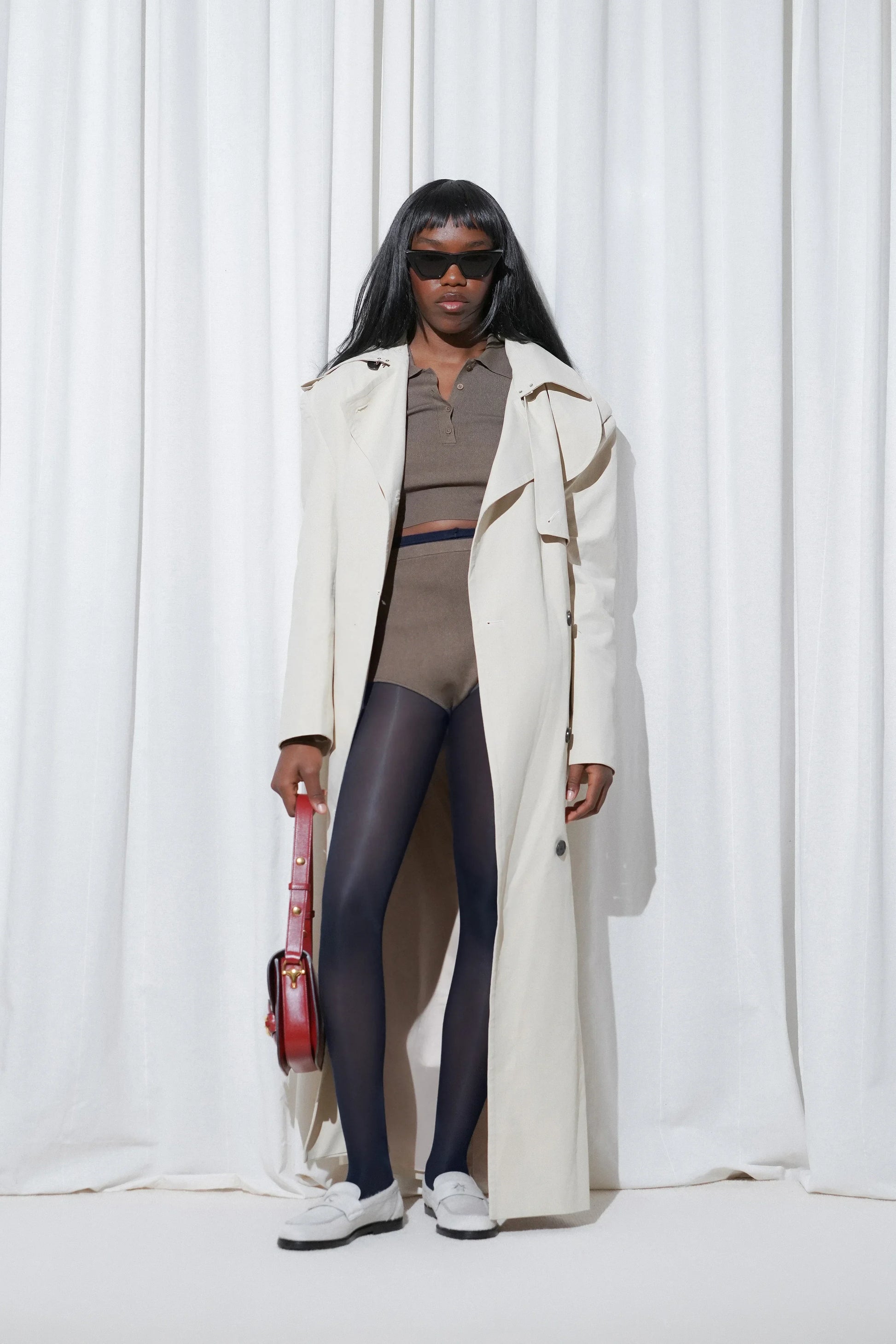 Model wearing a casual weekend outfit with a long cream trench coat, khaki knit crop top and shorts, EDLA Galaxy Navy Tights, and EDLA Jupiter White Loafers.