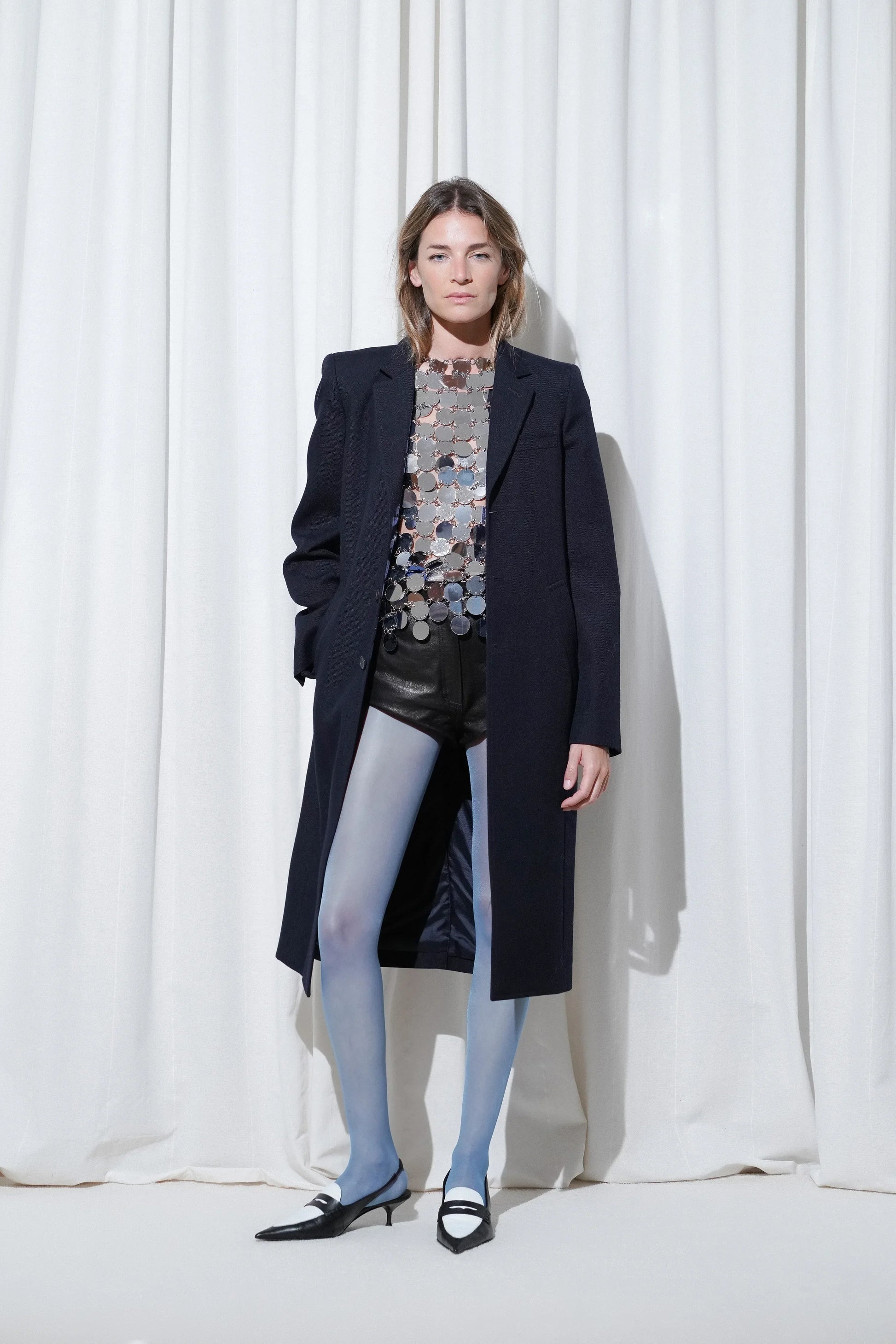 Model wearing a statement party look with a navy tailored coat, silver disc sequins embellished top, black leather shorts, EDLA Galaxy Blue Tights, and EDLA Uranus Black and White Slingbacks.