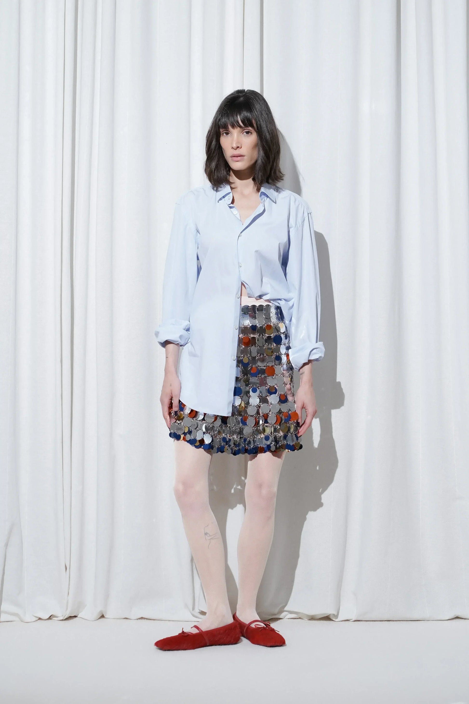 Model wearing a date outfit with an oversized light blue button-down shirt, a mini skirt made of mirrored paillettes, and EDLA Galaxy Pale Pink Tights and EDLA Mars Red Ballet Flats.