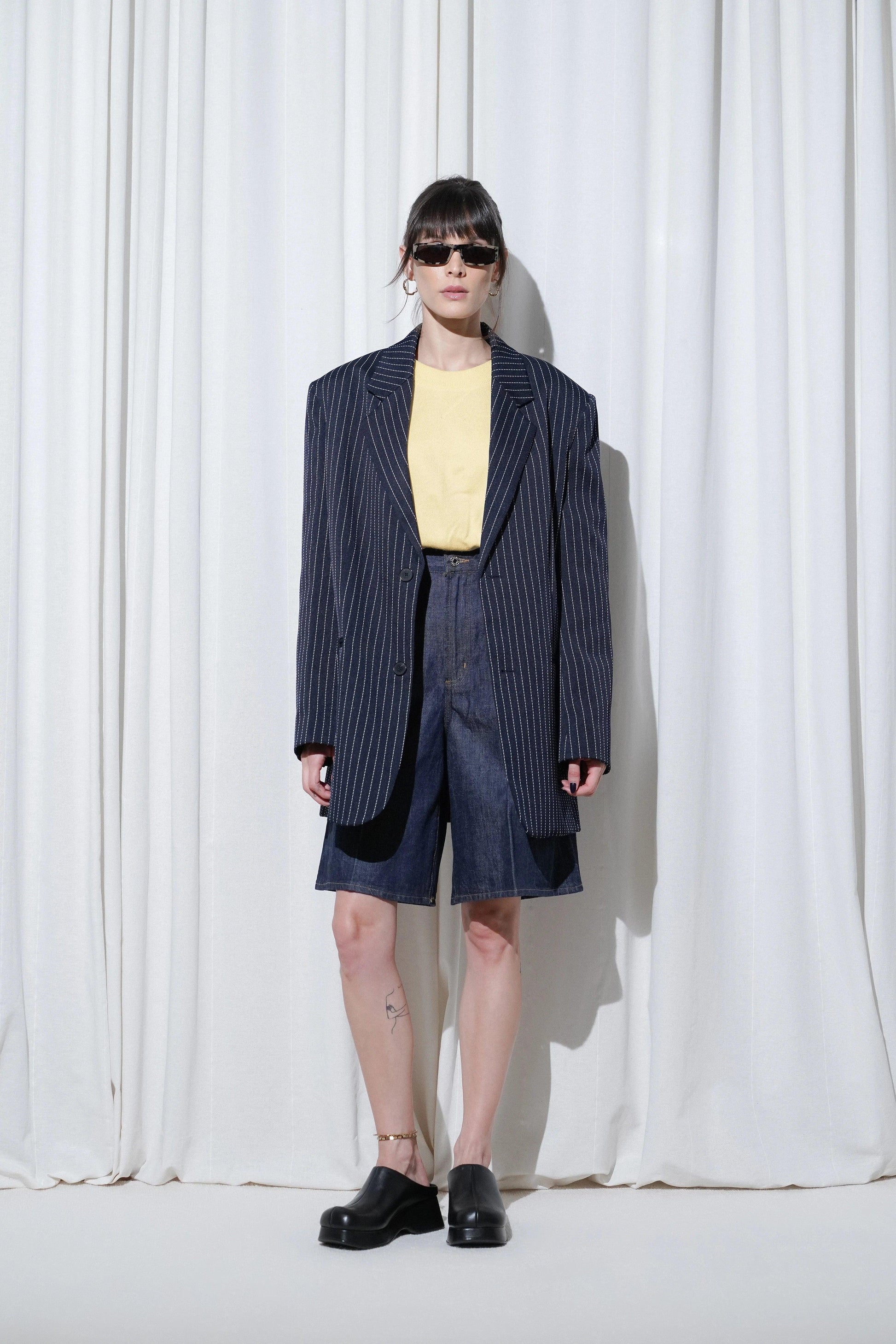 Model in a casual business outfit with pinstripe oversized blazer, yellow top, shorts, sunglasses, gold anklet and EDLA Venus Black Mules.