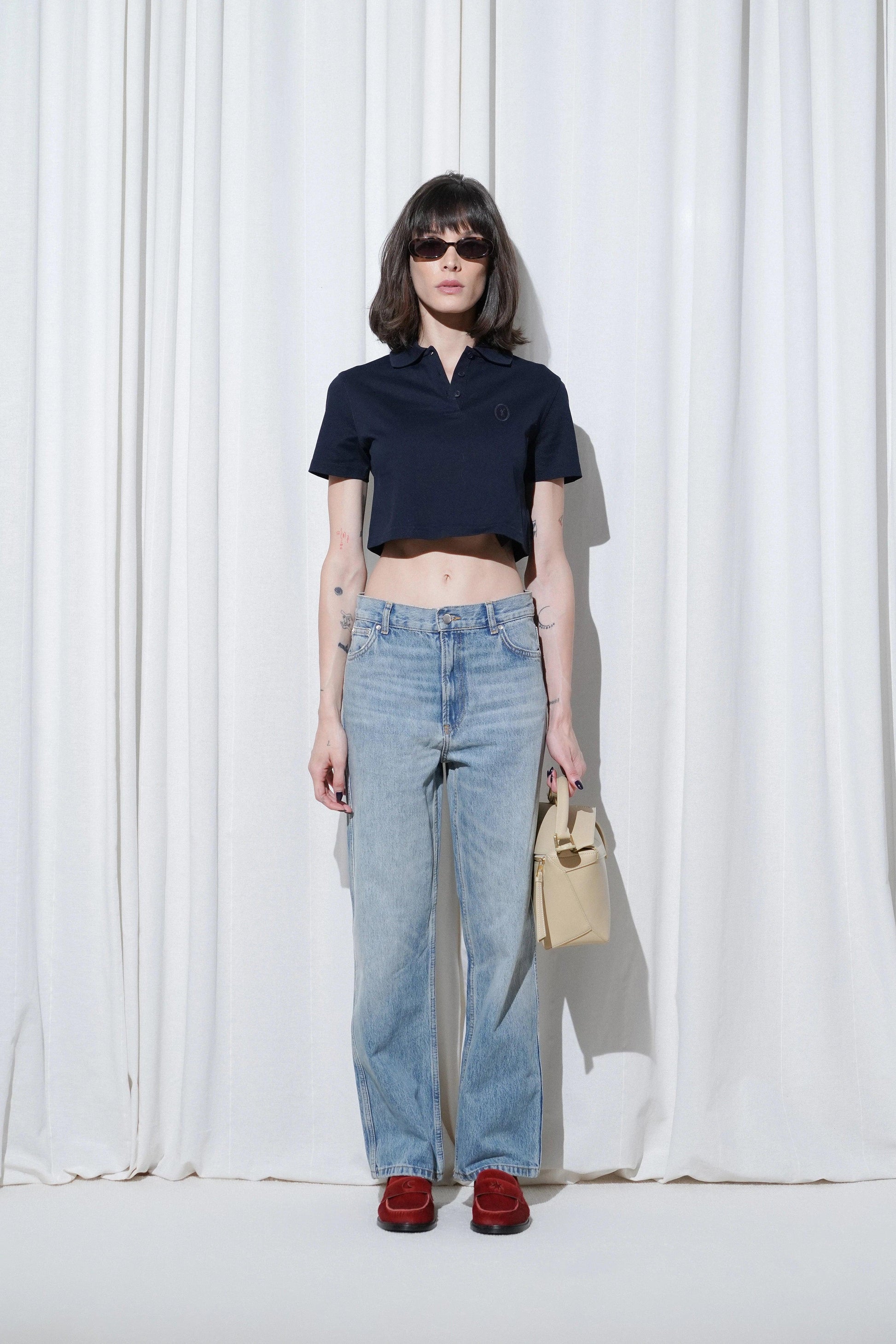 Model in a casual outfit with navy crop polo shirt, blue straight-leg jeans, beige handbag, and EDLA Jupiter Red Loafers.