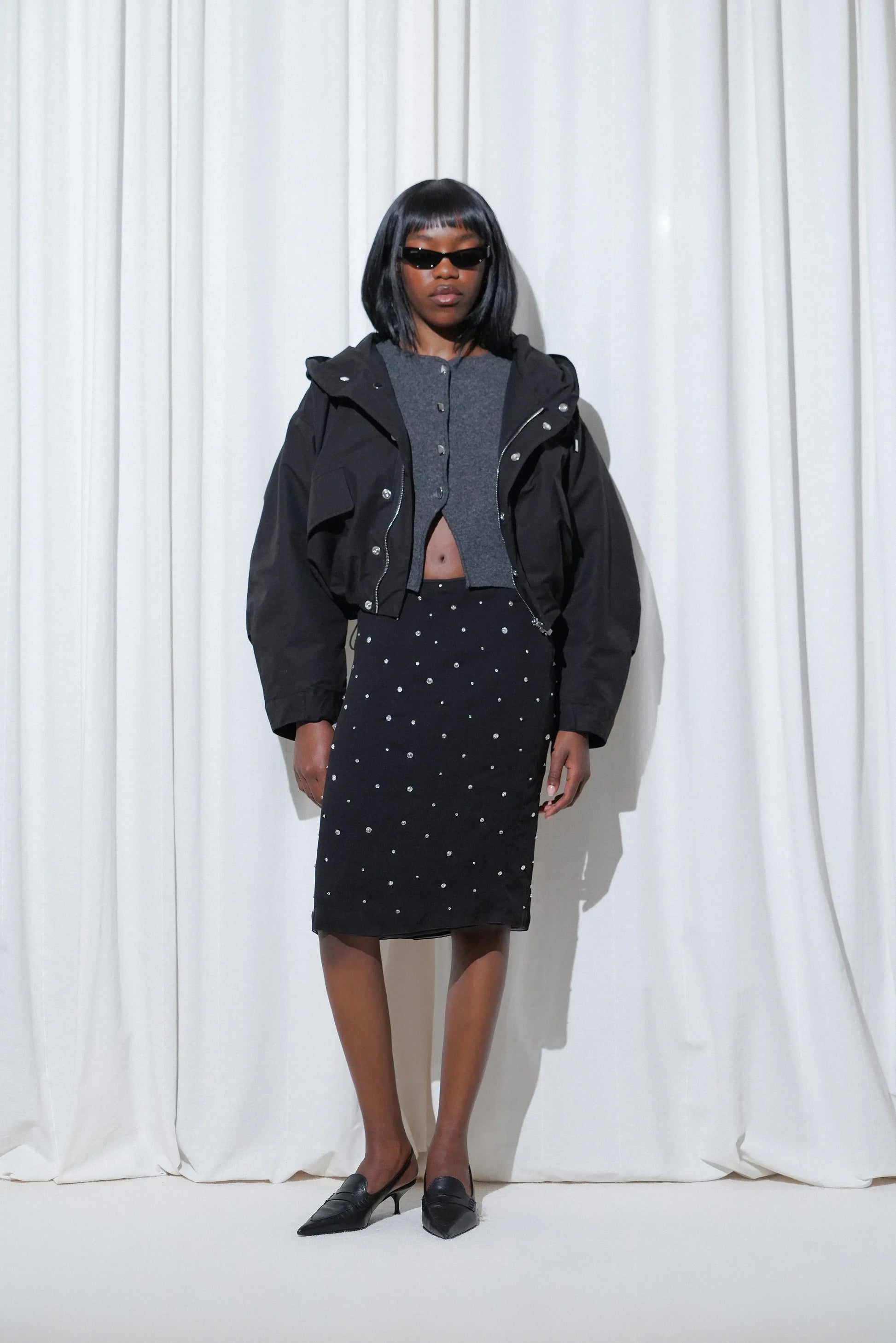 Model in a work outfit with cropped grey cardigan, crystal-embellished black midi skirt, cropped bomber jacket, and EDLA Uranus Black Slingbacks.